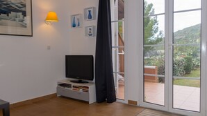 A living room with a flat-screen TV on a white entertainment unit, a wall-mounted lamp, and a view of a garden through a sliding glass door. - T2 1 bedroom 4 people for rent 83700 Saint Raphaël / Le Dramont  (Saint-Raphaël)