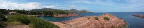 Un paisaje costero con terreno rocoso, mar azul cristalino y una colina distante con edificios.