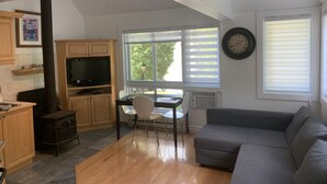 A living room with a gray sofa, a wooden dining table with chairs, a TV, and a kitchen area with cabinets and a stove. - Perfect Location / Perfectly Located Ski-in / Ski-out  (Mont-Tremblant)
