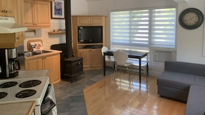 A compact kitchen with a stove, sink, and a black wood-burning stove. There is a dining area with a table and chairs, and a gray sofa in the living room. - Perfect Location / Perfectly Located Ski-in / Ski-out  (Mont-Tremblant)