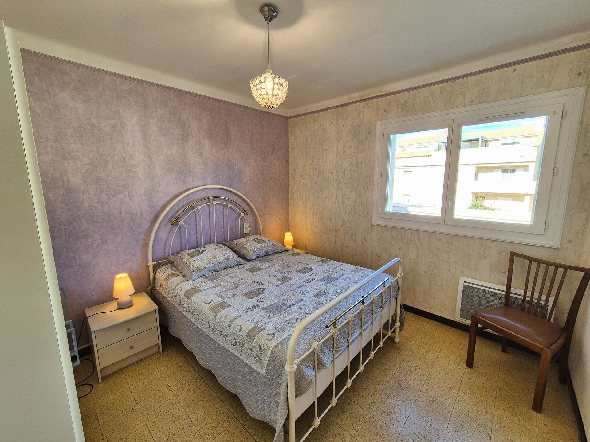 Une chambre à coucher avec un lit en métal, une chaise en bois, une table de chevet avec une lampe et une fenêtre donnant sur des immeubles.