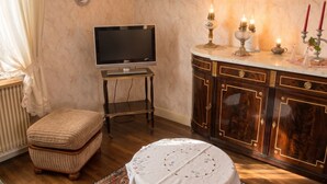 A room with a television on a stand, a cabinet with decorative items, a small table with a tablecloth, and a decorative wall piece. - Homerez - Apartment in Kientzheim (Kientzheim)