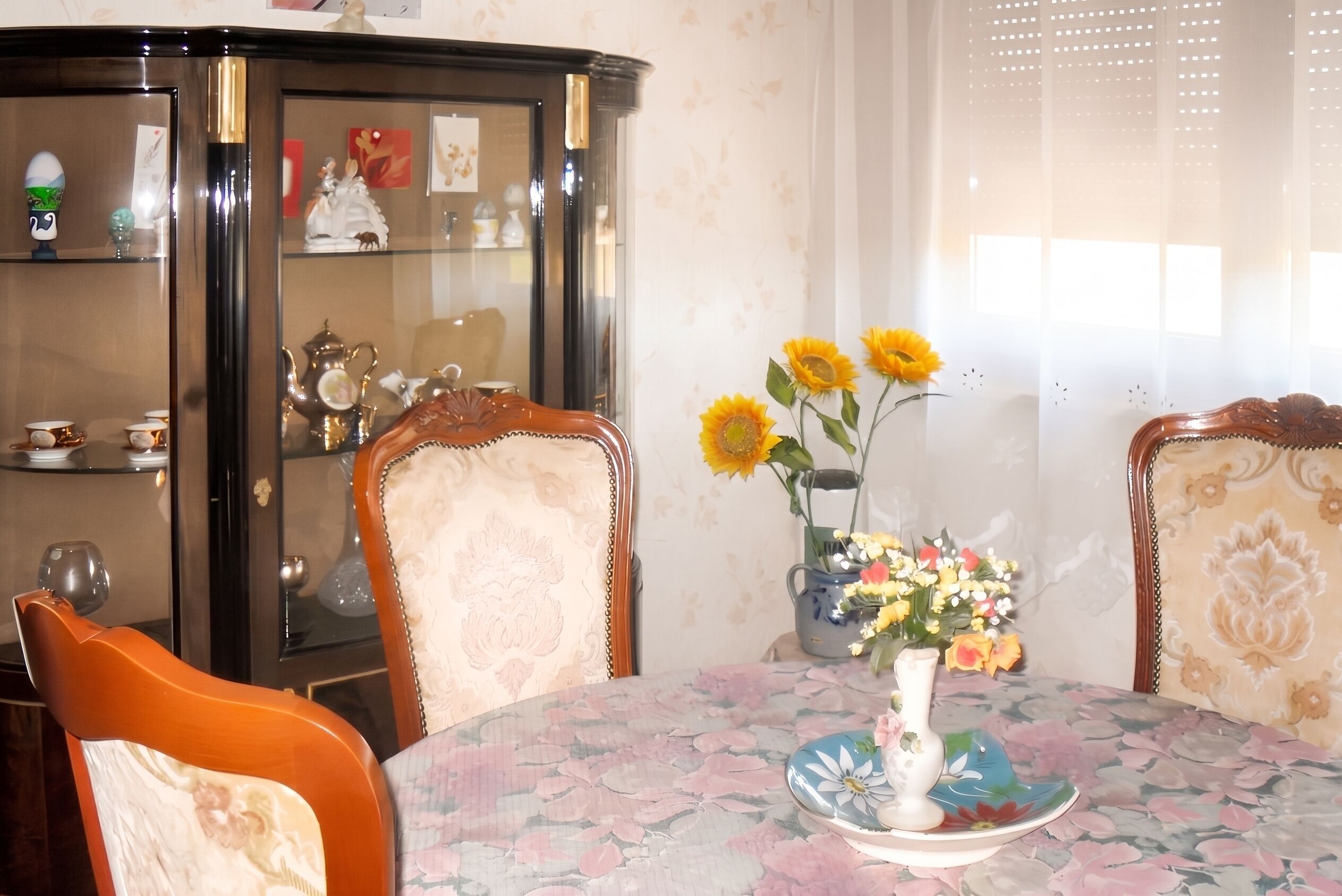 A dining room with a floral tablecloth, a vase with sunflowers, and a cabinet with glass doors.