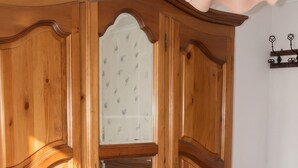 A wooden wardrobe with mirrored doors, a wicker stool, and a bed with a floral bedspread. - Homerez - Apartment in Kientzheim (Kientzheim)