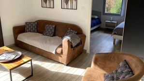 A modern living room with a brown sofa, a wooden coffee table, and a brown armchair. There are two framed artworks on the wall, a bed with a blue bedspread, and a window with a view of greenery.
