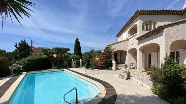 Una piscina con bordo in pietra, circondata da un giardino con diverse piante e un edificio con archi e tetto piastrellato.