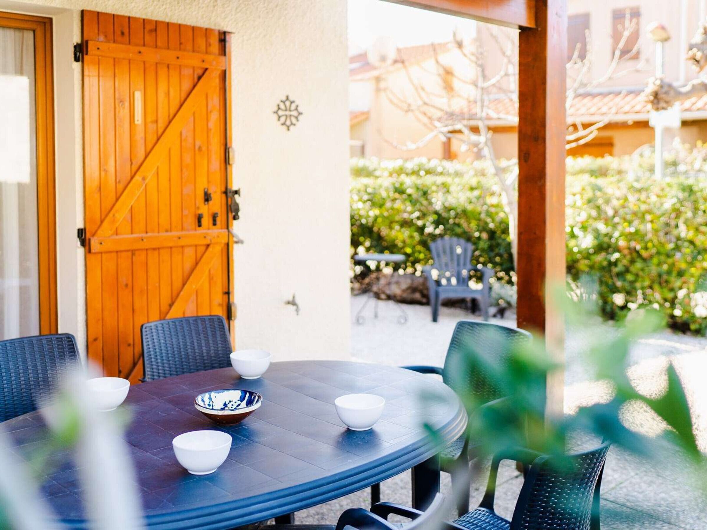 A round table with chairs and bowls, a wooden door, and a view of a garden with a chair and table.