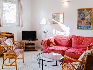 Un salon avec un canapé rouge, des fauteuils en osier, une table basse en verre, un téléviseur posé sur un meuble en bois et une lampe sur pied.