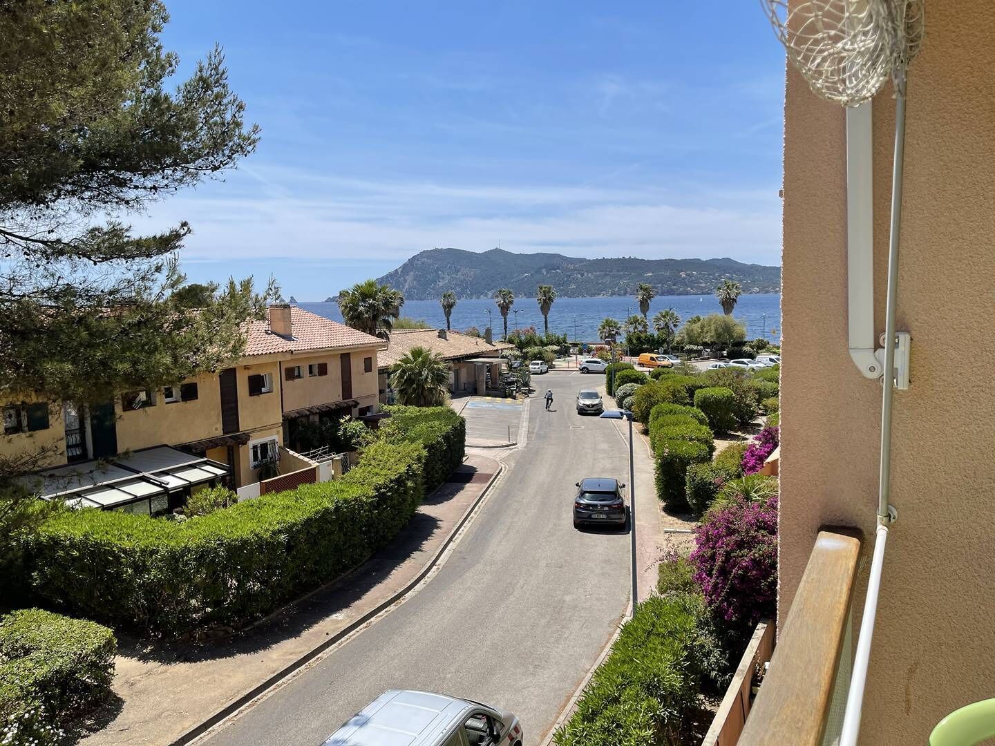 A residential street with houses, parked cars, and a clear view of the sea and mountains in the background.