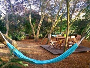 A hammock between trees, a wooden deck, and a table with chairs in a wooded area.