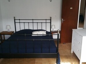 Ein Schlafzimmer mit einem Metallbettgestell, einer dunkelblauen Matratze, zwei Nachttischen mit Lampen und einer weißen Kommode.