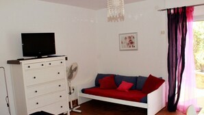 A bedroom with a bed, a white dresser, a television on top, a fan, a sofa with red cushions, and a window with curtains. - Studio & large garden 200m Calvi beach (Calvi)