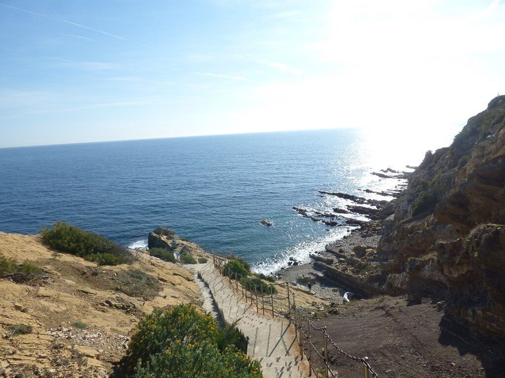 A coastal landscape with a rocky cliff, a sea view, and a paved path leading down to the water.