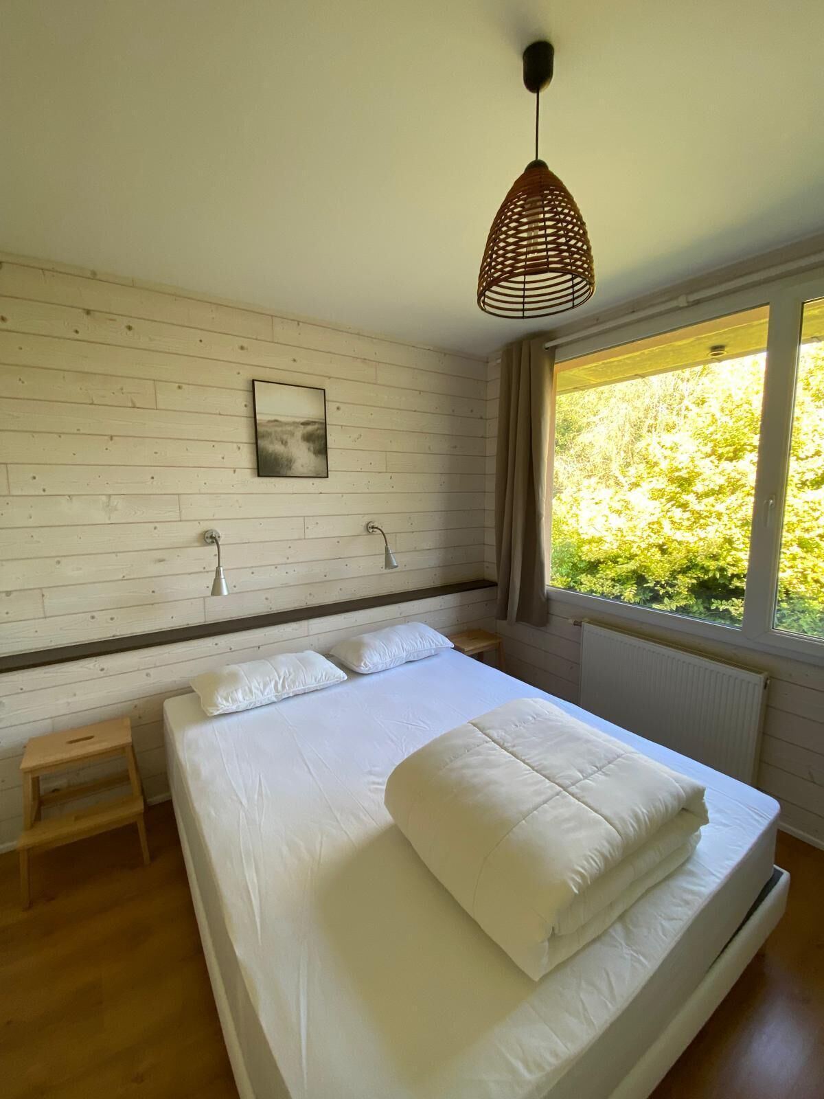 Une chambre avec un grand lit, des murs en bois, une fenêtre donnant sur un paysage verdoyant et un luminaire suspendu.