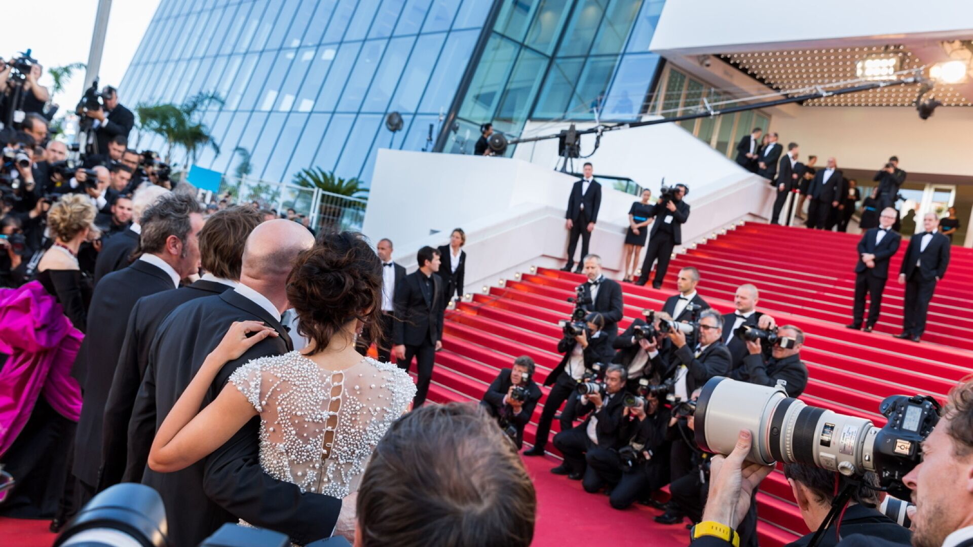 A red carpet event with people in formal attire, a modern building with glass windows, and photographers capturing the event.