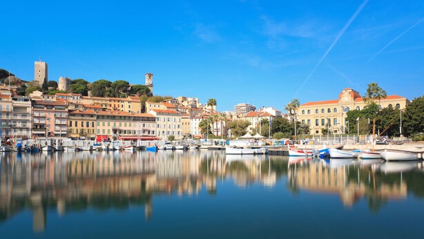 Une ville côtière dotée de bâtiments historiques, d’un port de plaisance avec des bateaux et d’un imposant beffroi.