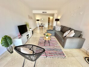 A modern living room with a grey sofa, a black chair, a TV, and a dining area in the background.