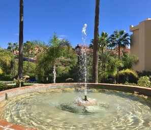 A fountain in a circular pool surrounded by palm trees and a building in the background.