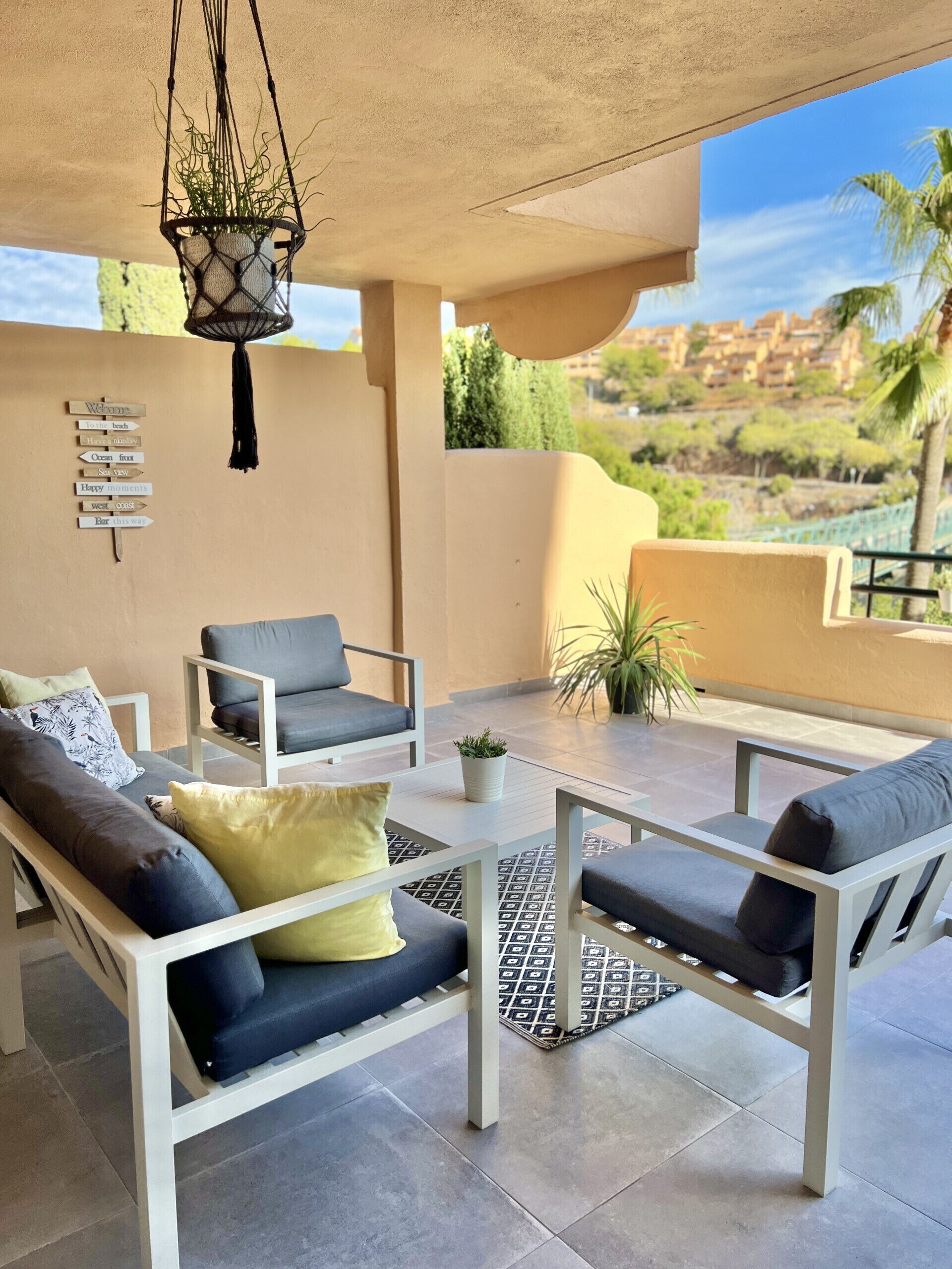 A covered outdoor seating area with two sofas and a table, a hanging plant, and a wall with directional signs.