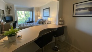 Een moderne hotelkamer met een gebogen bureau, twee barkrukken, een groot raam met uitzicht, een flatscreen televisie en een potplant.
