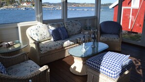Una habitación iluminada por el sol con muebles de mimbre, una mesa de vidrio y vistas al agua y edificios.