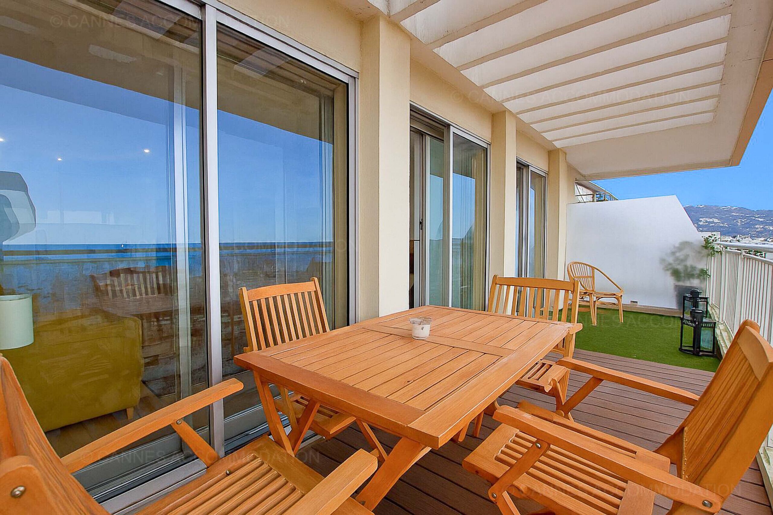 A wooden deck with a table and chairs, large sliding glass doors, and a view of the sea.