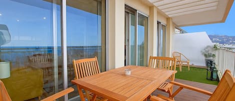 A wooden deck with a table and chairs, large sliding glass doors, and a view of the sea.