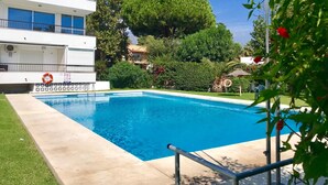 Una piscina con acqua cristallina circondata da un solarium in cemento. Si vede un edificio bianco con balconi e un albero sullo sfondo.