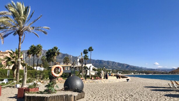 Una spiaggia con palme, un salvagente e una grande sfera nera. In secondo piano, persone e sedie a sdraio.