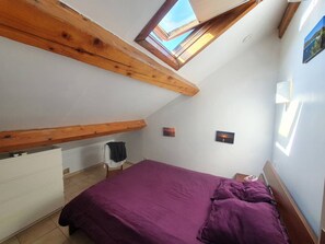 A bedroom with a sloped ceiling, wooden beams, a bed with purple bedding, a white dresser, and two small paintings on the walls. - Beachfront, Comfortable, AIR CONDITIONER (ST MANDRIER SUR MER)