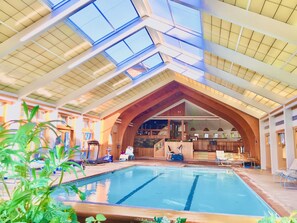An indoor swimming pool with a high, sloped ceiling and skylights, surrounded by wooden beams and a bar area.
