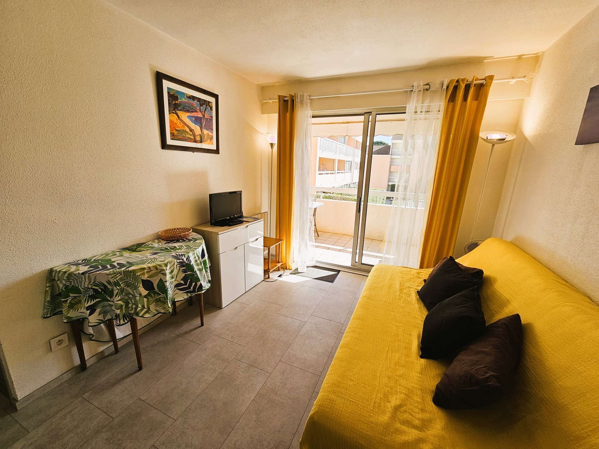 A small, bright room with a bed, a desk, and a TV. There is a window with curtains and a view of buildings outside.