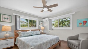 Une chambre à coucher comprenant un lit, des tables de chevet, une chaise, un ventilateur de plafond et des fenêtres donnant sur un paysage verdoyant.
