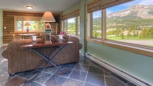 Una habitación acogedora con sofá, mesa y lámpara. Amplias ventanas ofrecen vistas a montañas y vegetación.