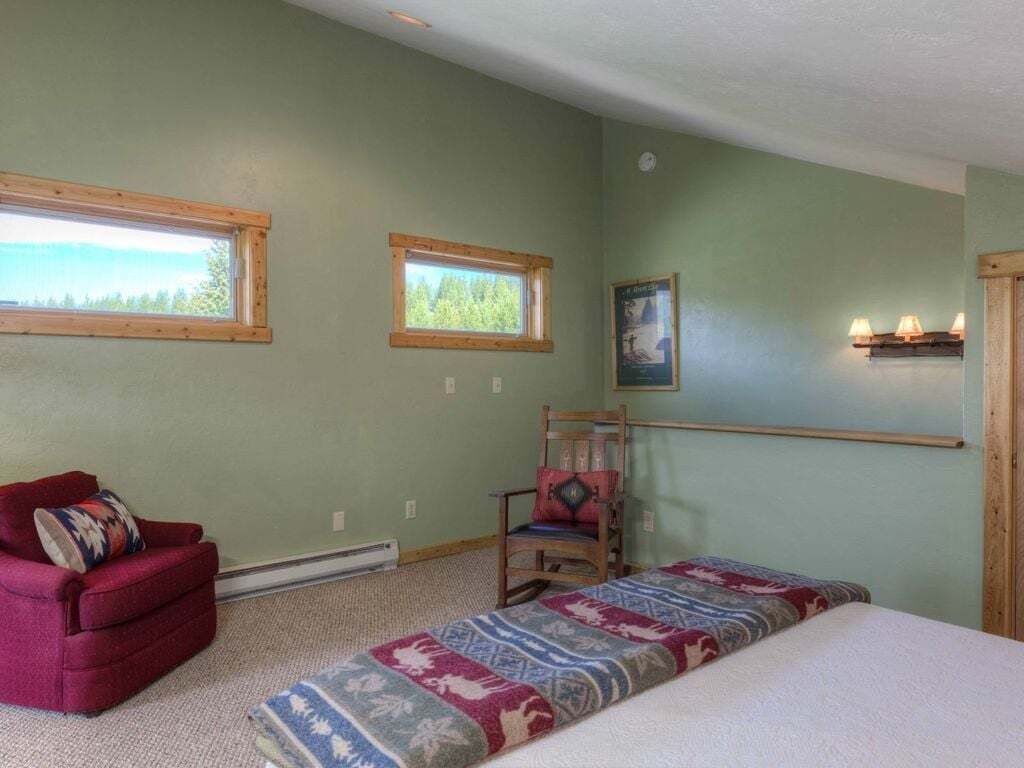 A room with a bed, a red armchair, a wooden chair, two windows, and a wall-mounted shelf with lamps.