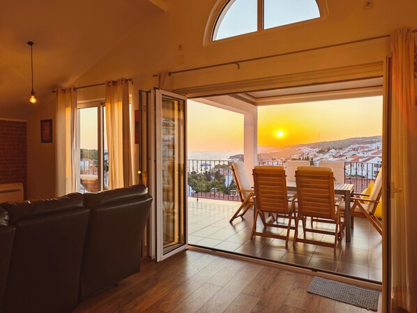 A room with a sofa, a dining table with chairs, and a view of the sunset over the sea.
