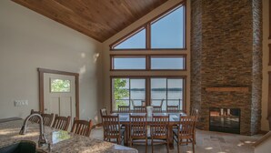 A spacious dining area with a large table and chairs, a stone fireplace, and large windows overlooking a body of water.
