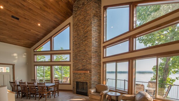 A spacious room with a high ceiling, large windows, a stone fireplace, and a view of a lake.