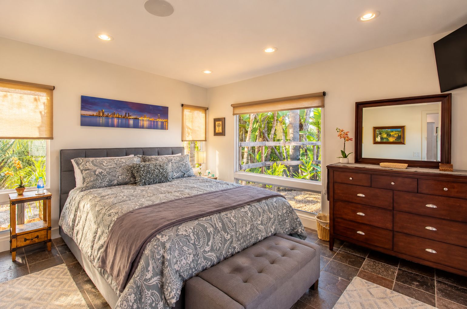 A bedroom with a large bed, a dresser, a window with a view of greenery, and a painting on the wall.