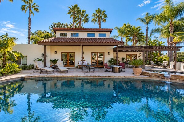 A poolside area with lounge chairs, a pergola, and a house with a tiled roof.