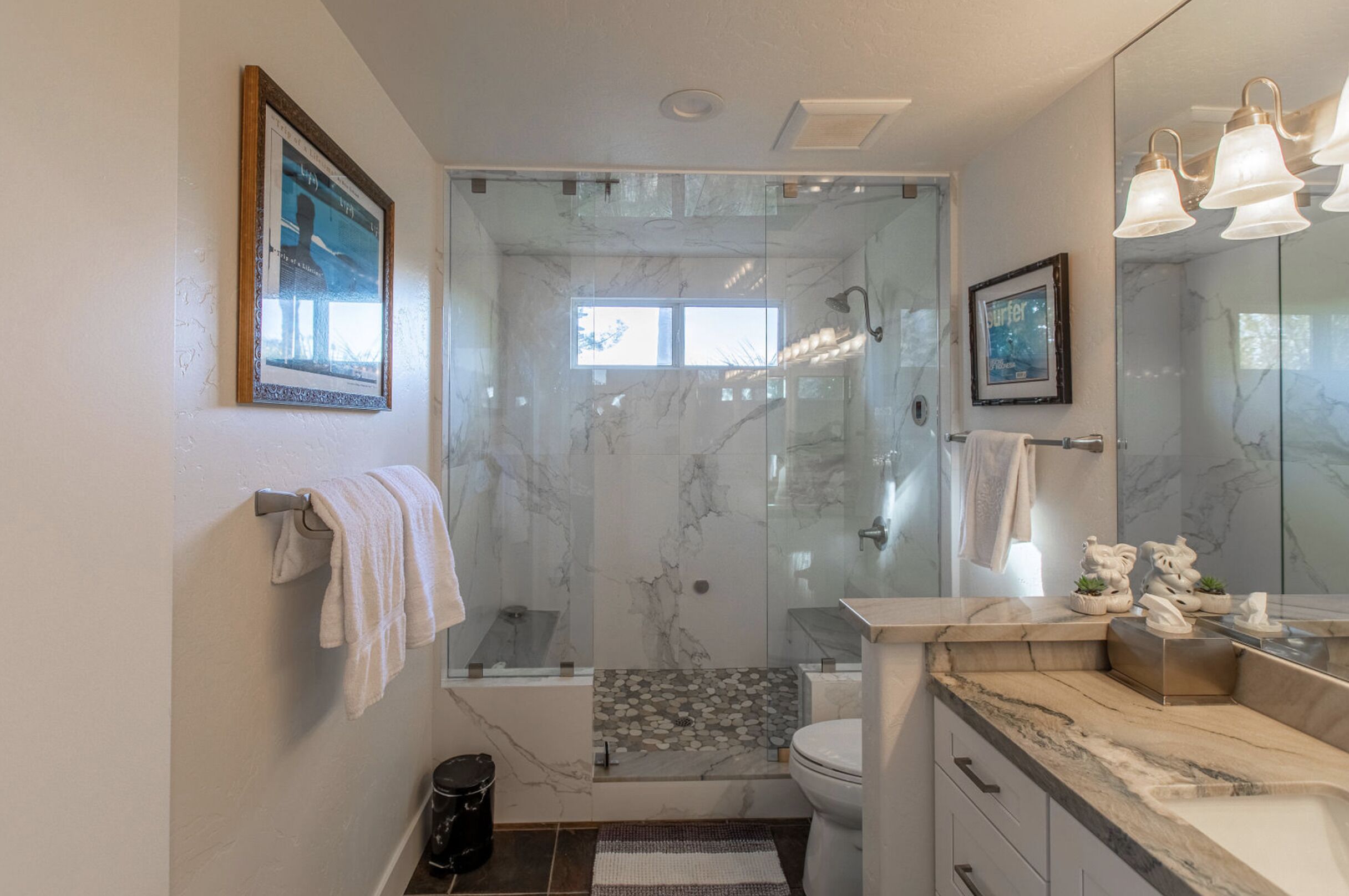 A modern bathroom with a glass-enclosed shower, marble walls, a toilet, and a vanity with a mirror and lights.