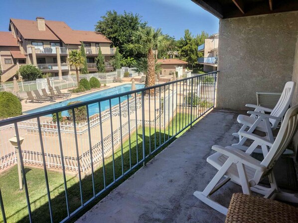 Un balcón con sillones blancos, una piscina y vistas a otros edificios y vegetación.