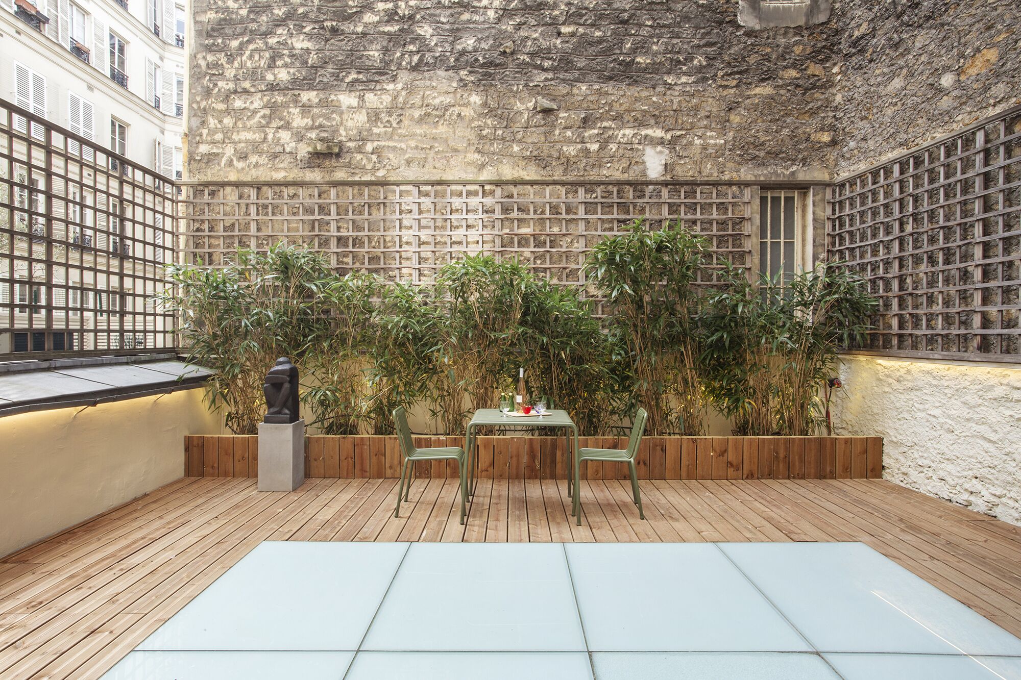 Una terraza de madera con una mesa y sillas de vidrio, rodeada de abundante follaje y un muro de ladrillo.