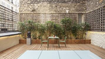 Una terraza de madera con una mesa y sillas de vidrio, rodeada de abundante follaje y un muro de ladrillo.