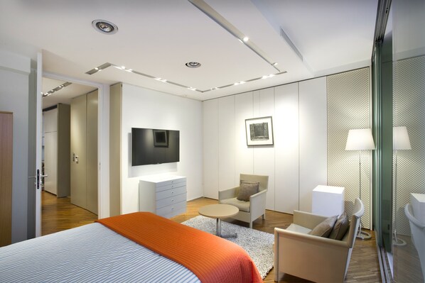 Ein modernes Hotelzimmer mit Bett, Fernseher, einer Sitzecke mit Sessel und Tisch sowie einem kleinen Schreibtisch.