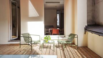 Un pequeño patio con dos sillas de metal verde y una mesa de vidrio, suelo de madera y vista hacia la habitación contigua.