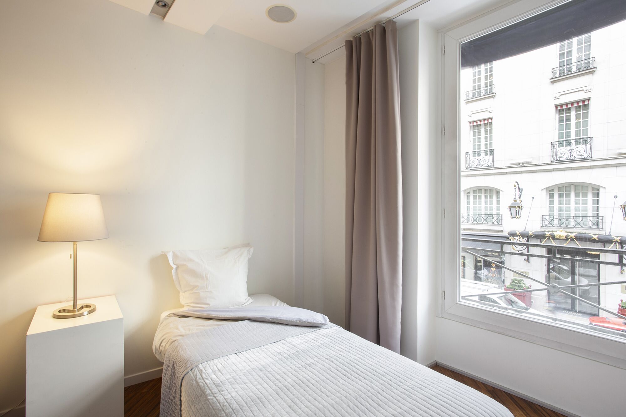 Ein Hotelzimmer mit Bett, einer Nachttischlampe, einem Fenster mit Vorhängen und Blick auf ein Gebäude mit Balkonen.