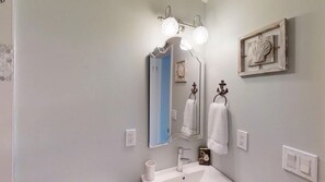 Une salle de bain avec un lavabo blanc, un miroir avec éclairage, une décoration en forme de coquillage encadrée, et une porte bleue en arrière-plan.
