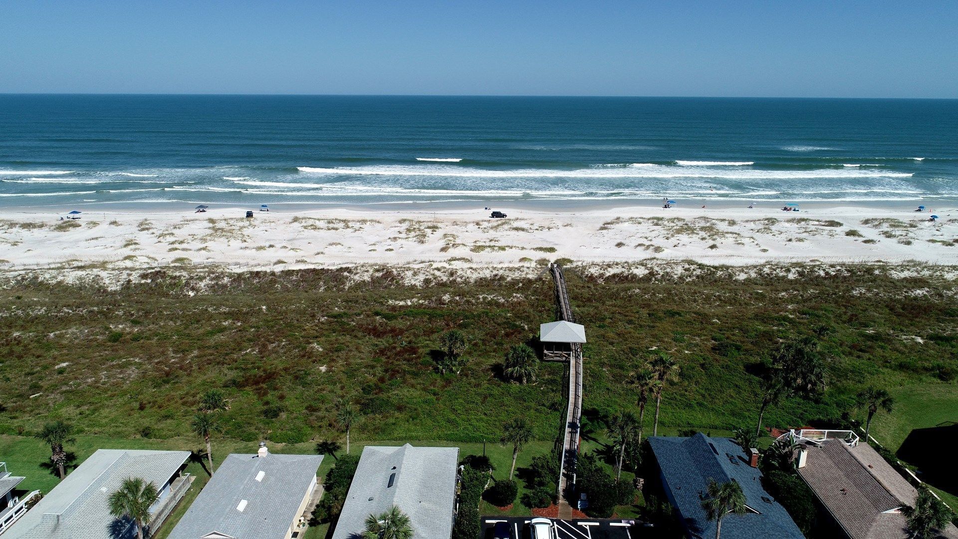 Une vue aérienne d’une plage, avec une vue dégagée sur l’océan, une plage de sable et quelques bâtiments aux toits plats.