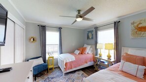Une chambre d’hôtel avec deux lits, un ventilateur de plafond, une petite table avec une lampe et une vue sur la mer grâce à de grandes fenêtres.